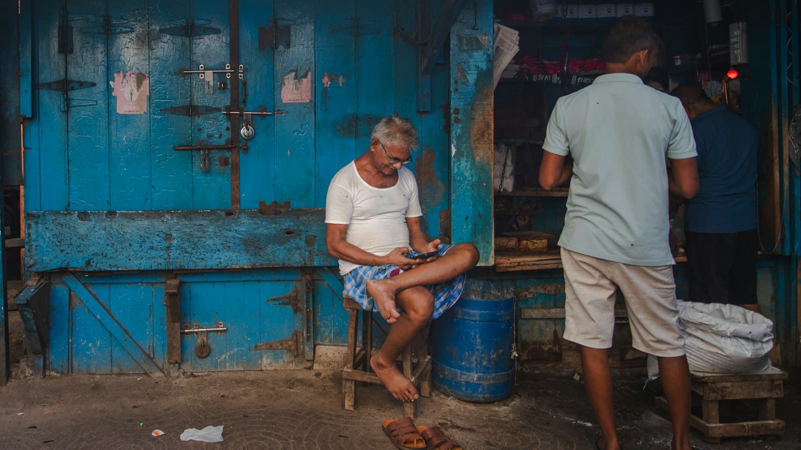 A man in a white vest sits on a wooden stool outside a blue shopfront, typing on his phone