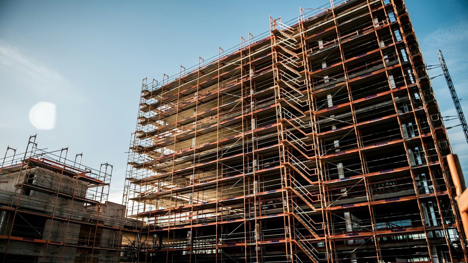 A multi-storey building wrapped in orange scaffolding against a blue sky, with a crane at the edge