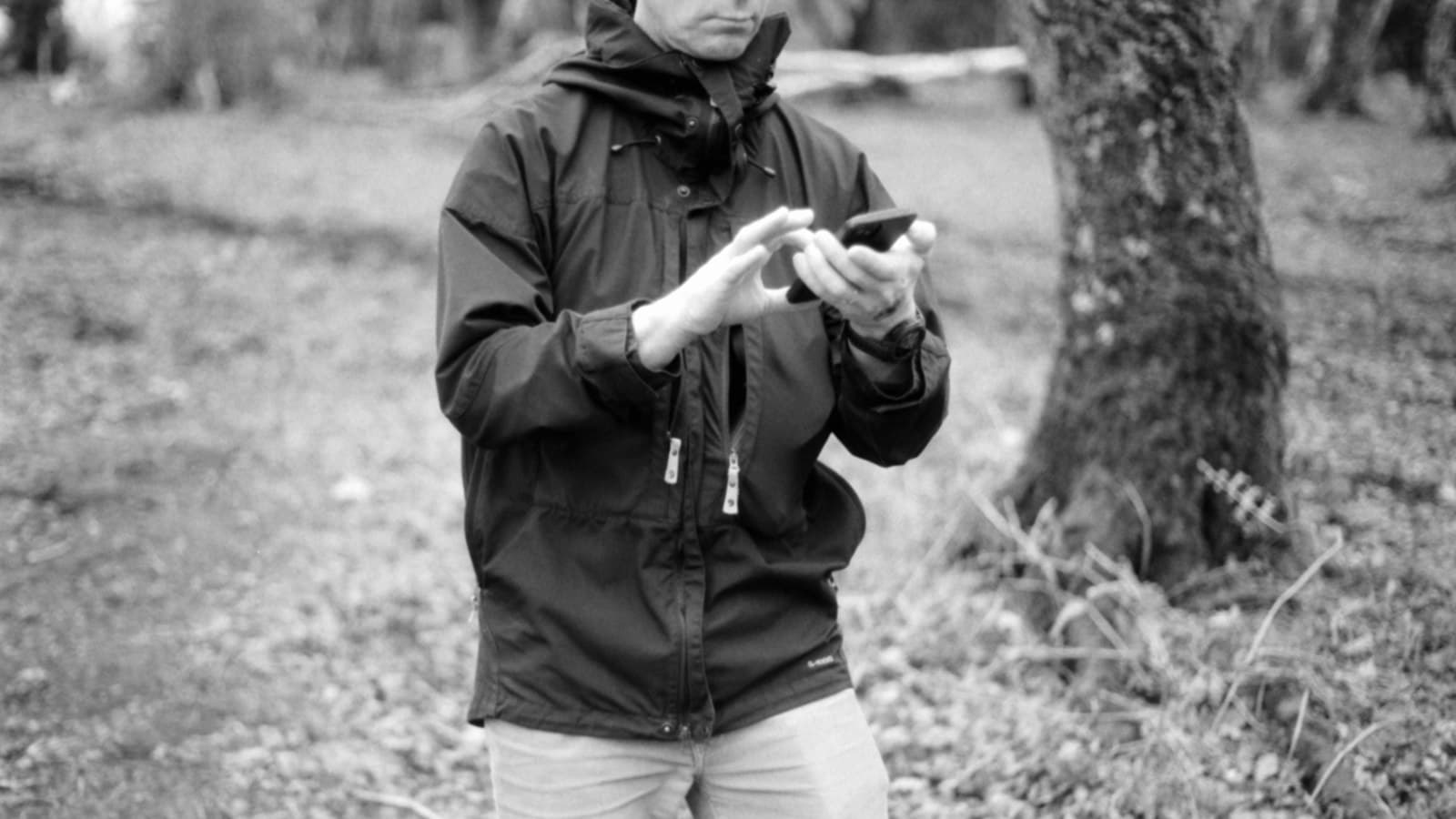 Black and white photo of a man in a rain jacket using a smartphone in a woodland clearing